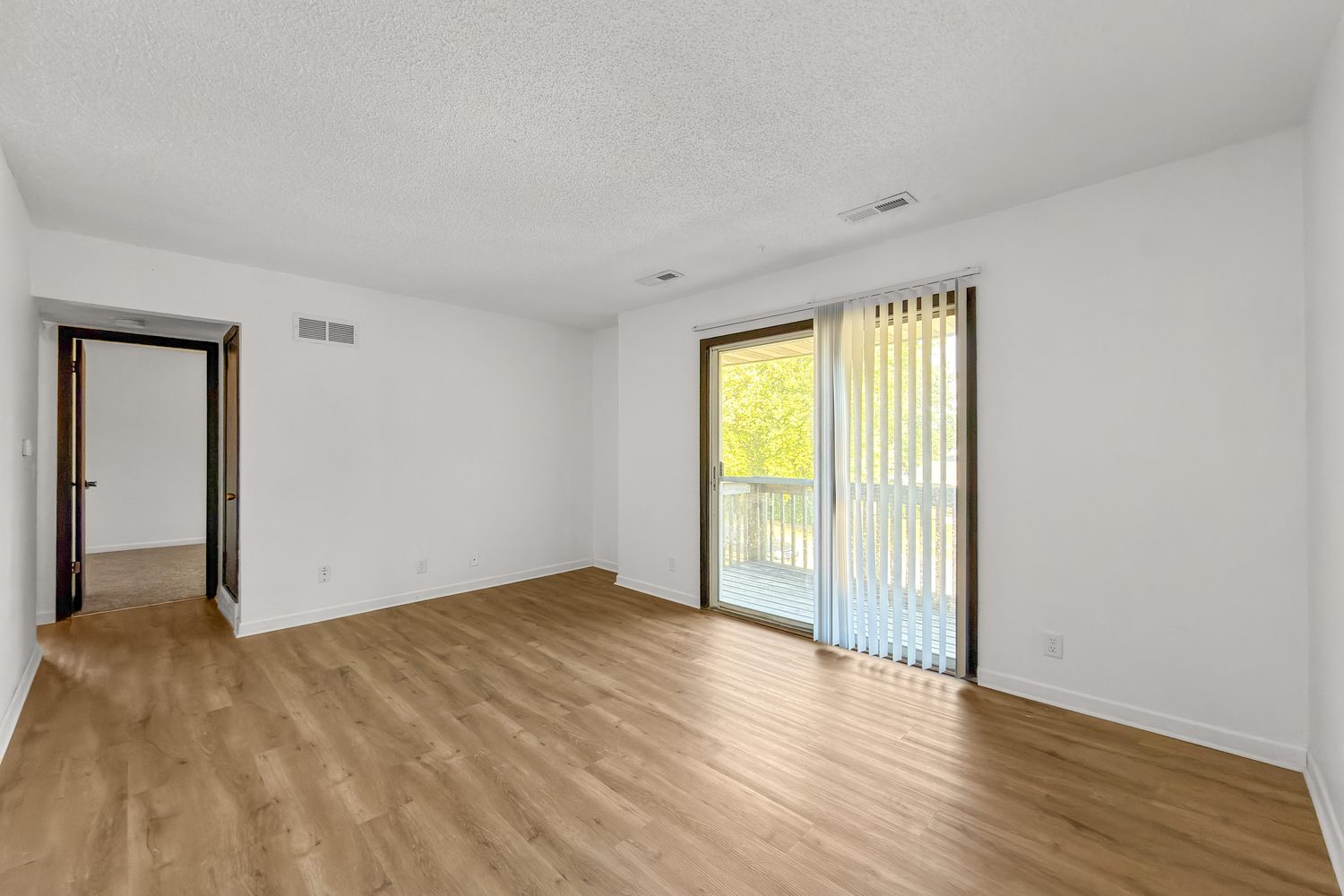 A living room with light wood-look plank flooring and a sliding glass door with vertical blinds opening to the balcony.
