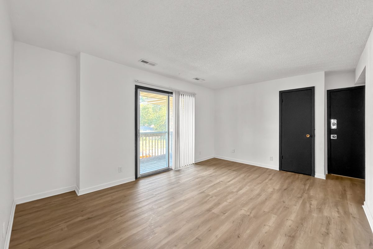 A living room with plank flooring, a sliding glass door to the balcony and the apartment entry door beside it.