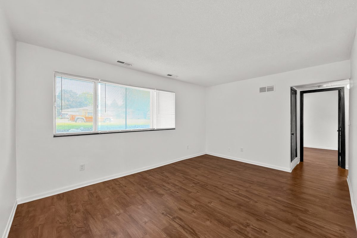 A living room with plank flooring, a wide window and the doorway to the bedroom hall.